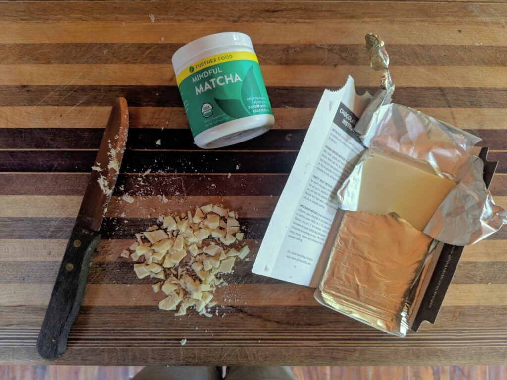 overhead image of an unwrapped white chocolate bar, chopped white chocolate pieces, and a container of Further Food Mindful Matcha